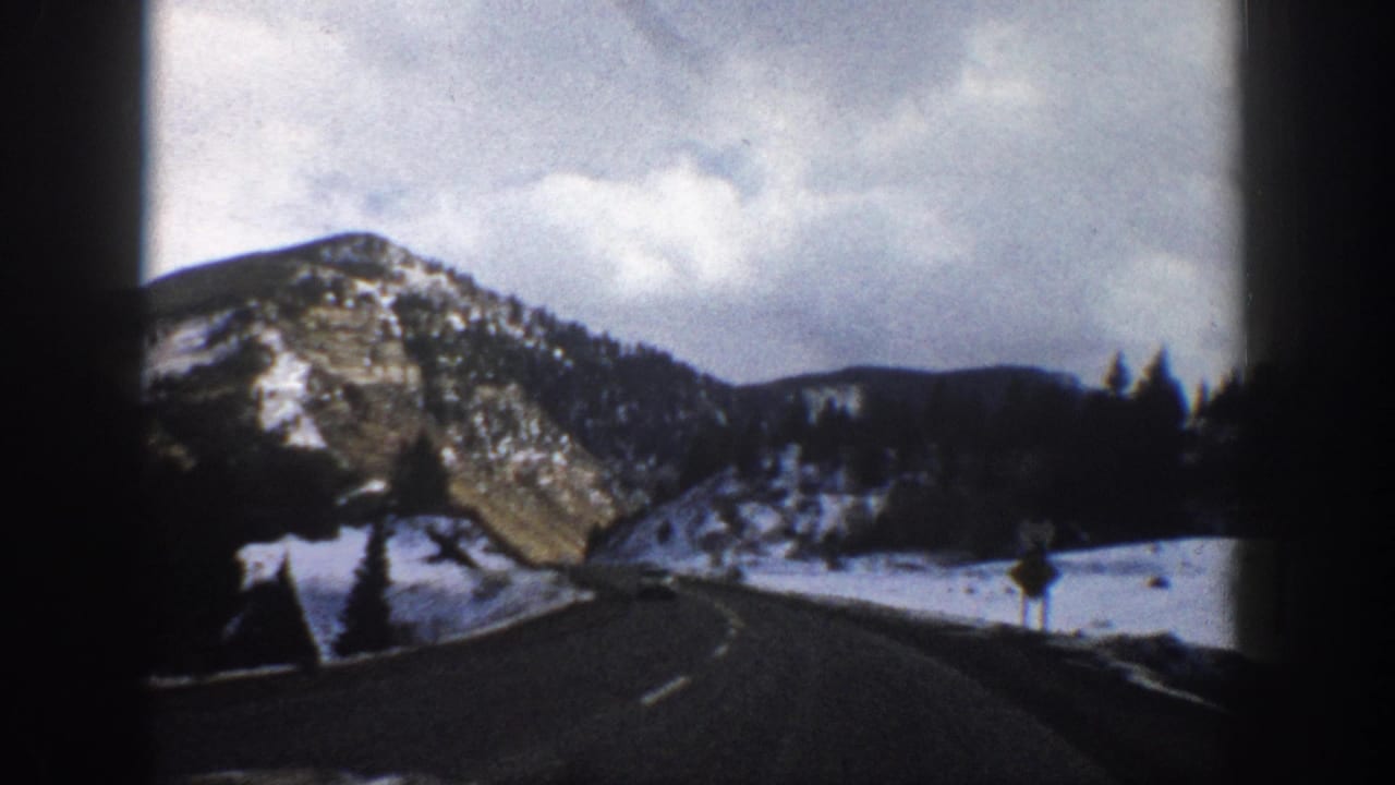 Curving mountain road bordered by snow, pine trees, and rocky hills.