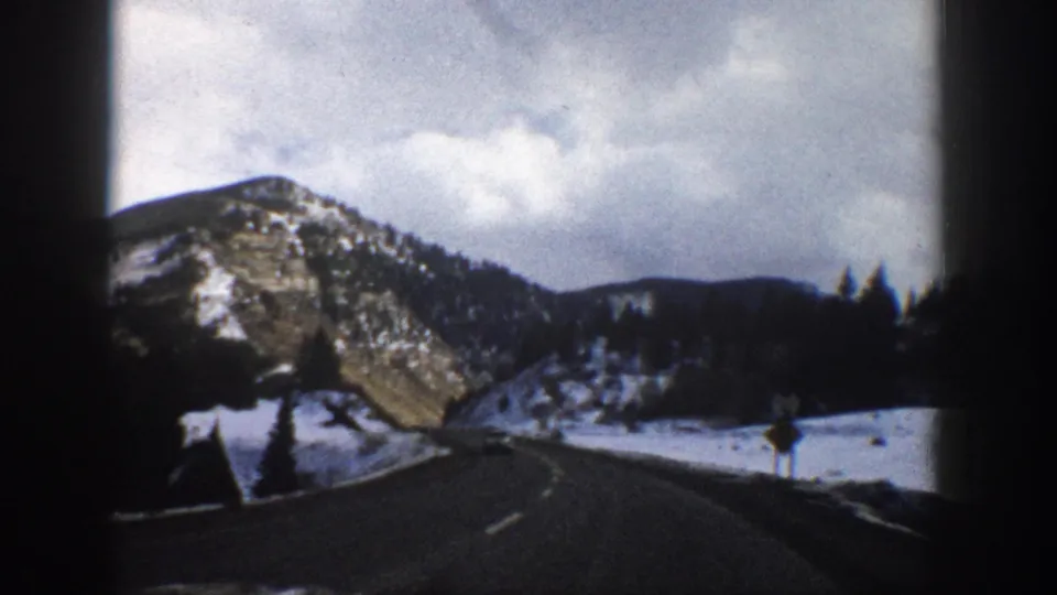 1961 Aspen, Colorado road-view frame with a winding asphalt road, snowbanks, dark pines, rocky slopes, and a small roadside sign.