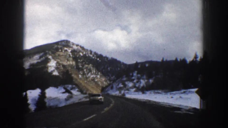 1961 Aspen-area 8mm road trip frame showing a winding road, snow-covered shoulders, pine-lined slopes, and a distant white car.