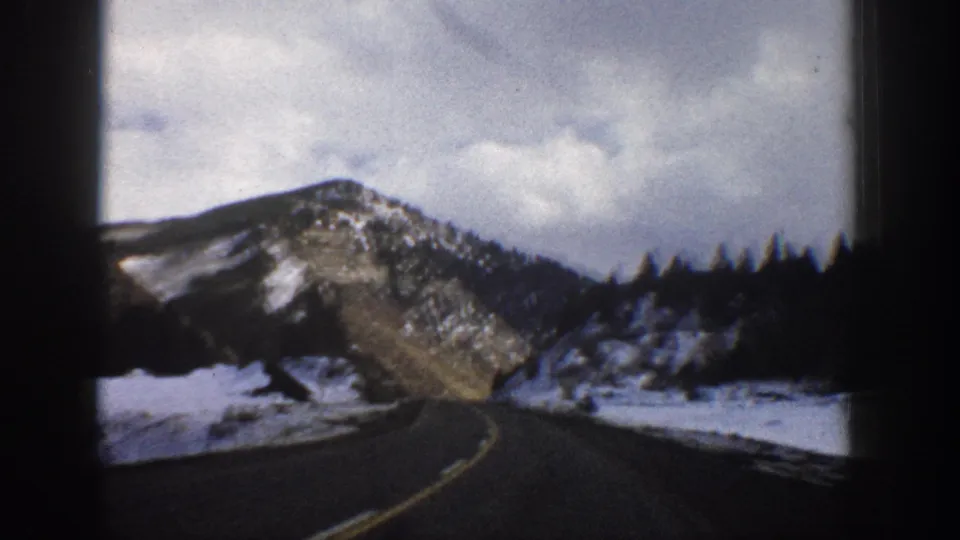 1961 Aspen, Colorado 8mm frame with a winding two-lane road, snow along the shoulder, pine trees, and a rocky mountain backdrop.