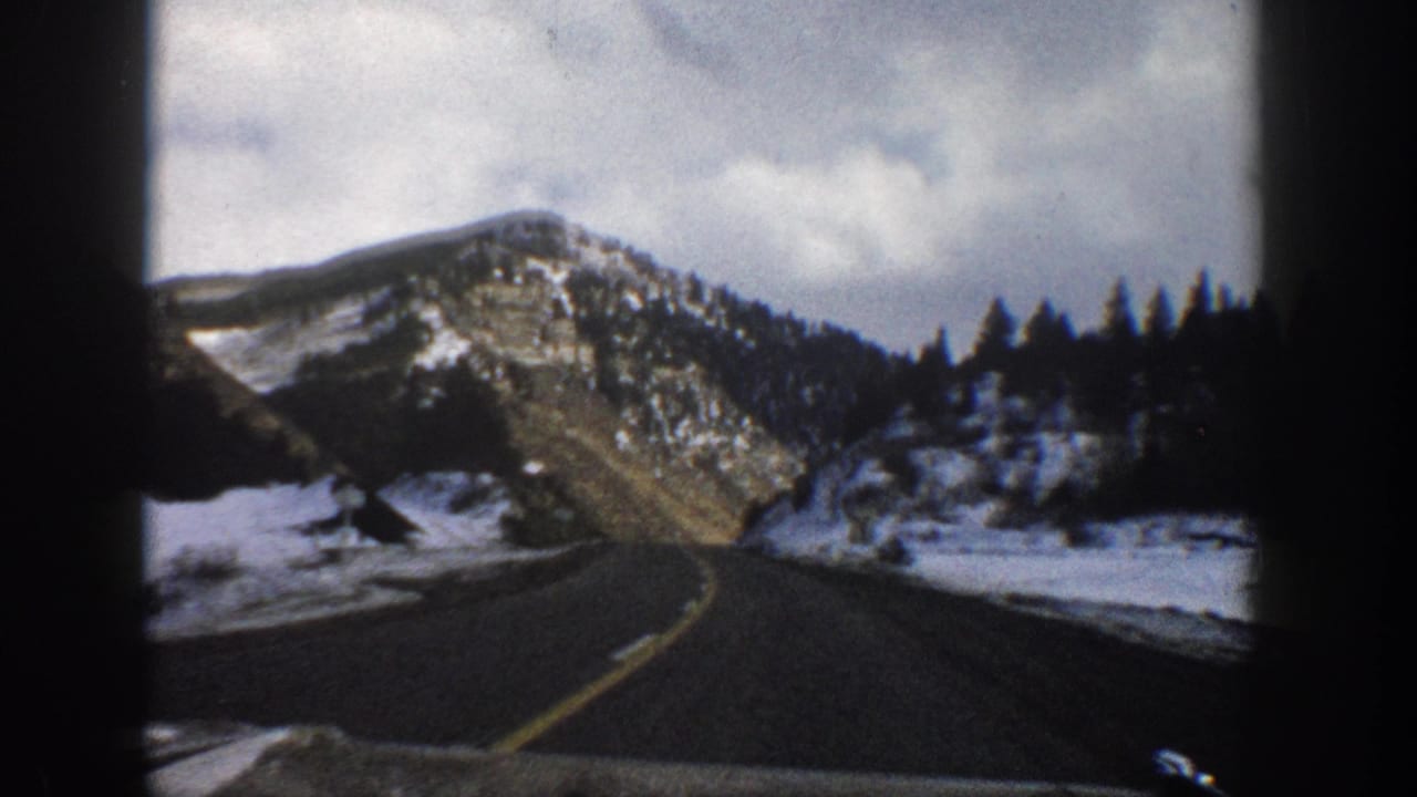 1961 Aspen, Colorado home-movie frame from inside a vehicle, looking along a curving highway toward snowy mountains and evergreens.