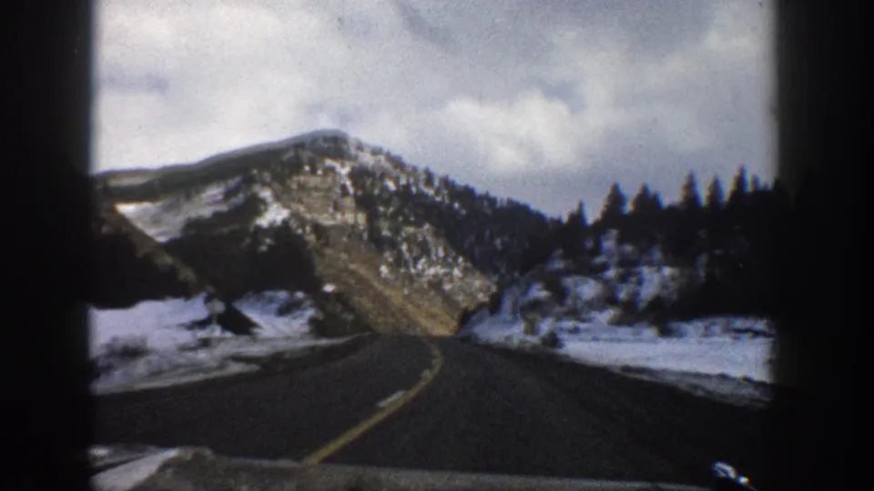 1961 Aspen, Colorado home-movie frame from inside a vehicle, looking along a curving highway toward snowy mountains and evergreens.