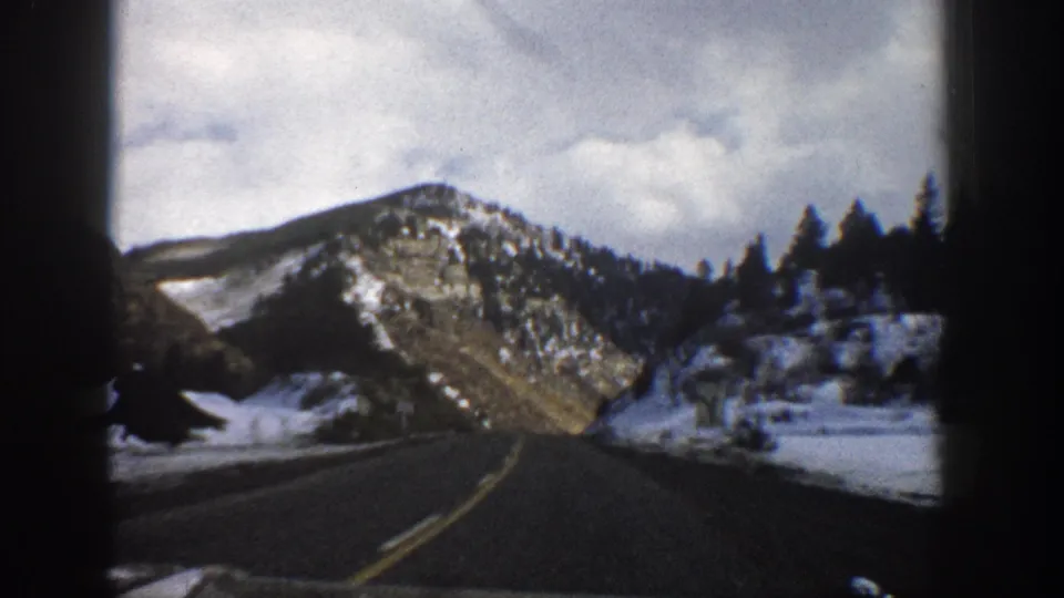 The moving-car viewpoint shows a winding Aspen-area highway bordered by snow, conifers, and a rocky mountain face in 1961 footage.