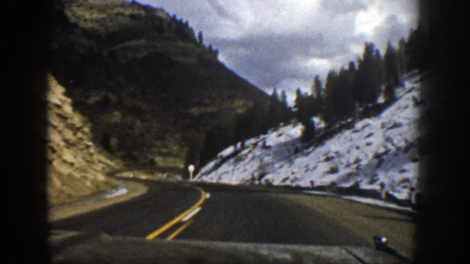 A 1961 Colorado mountain road scene shows the car approaching a bend, with snow on the right hillside and dark forested slopes ahead.