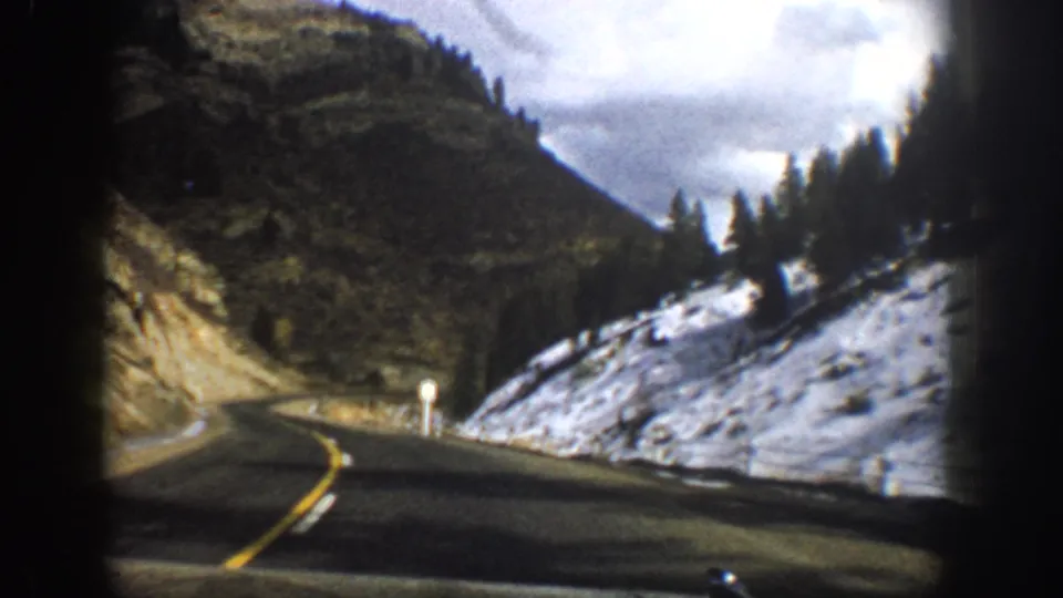 From inside the moving car, the Aspen-area highway bends beside snow-covered ground, a small roadside sign, and conifer-lined mountain slopes.