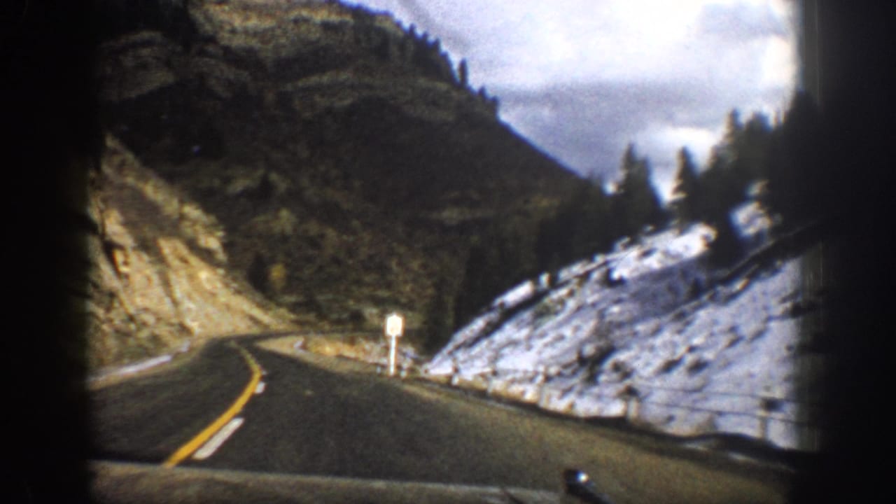 1961 Aspen-area home-movie view of a curving mountain road with snow patches, rocky hillside, trees, and cloudy sky.