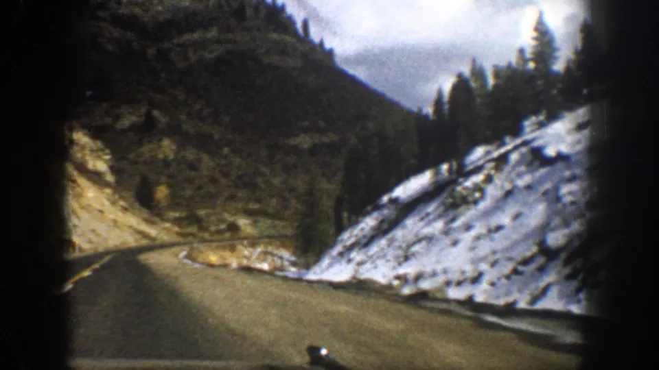 1961 Aspen-area traveler perspective on a curving road bordered by snow, evergreens, and a steep rocky hillside.