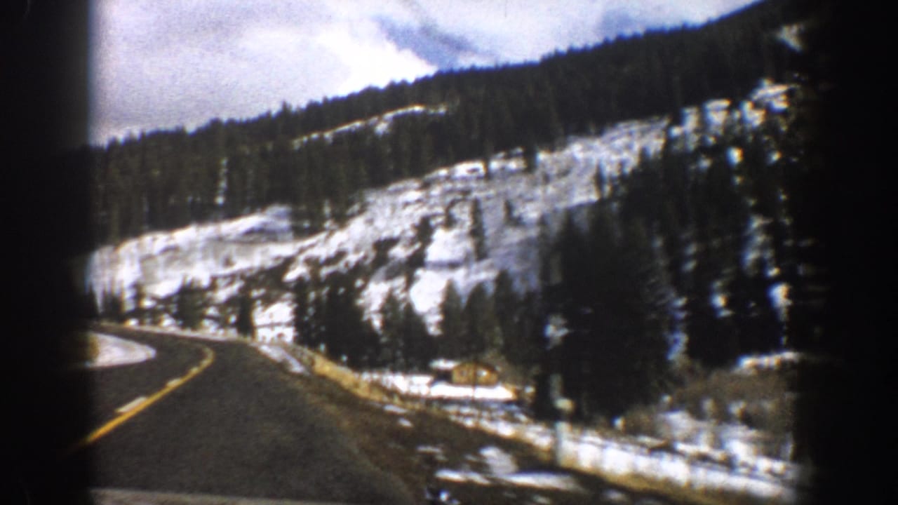 1961 Aspen, Colorado home-movie frame showing a curving road, snowbanks, evergreen trees, and a hillside cabin.