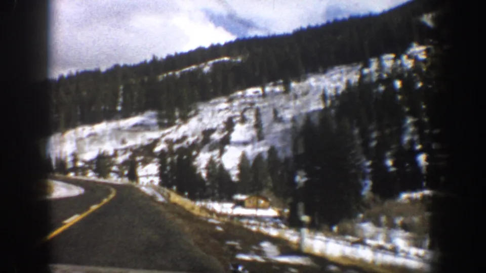 1961 Aspen, Colorado home-movie frame showing a curving road, snowbanks, evergreen trees, and a hillside cabin.