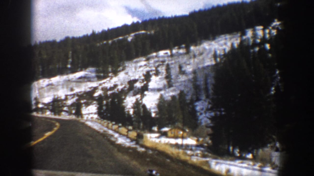 1961 Aspen, Colorado frame with a paved mountain road curving past snow, trees, roadside posts, and a small cabin.