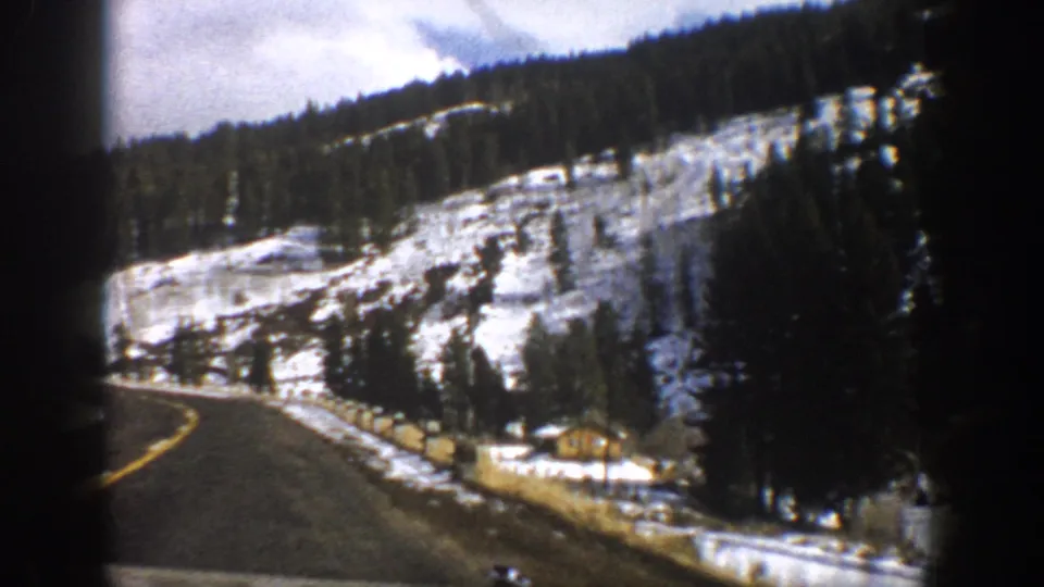 1961 Aspen, Colorado frame with a paved mountain road curving past snow, trees, roadside posts, and a small cabin.