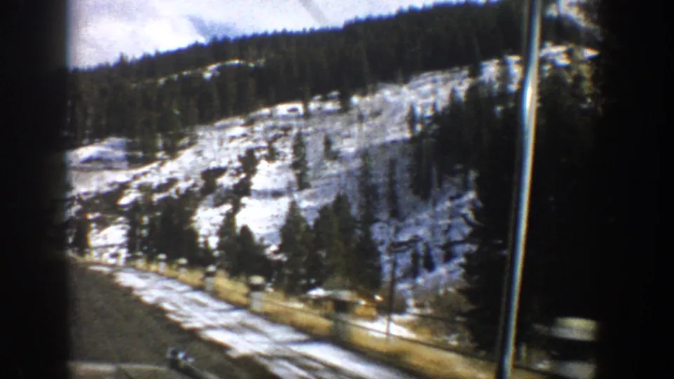 1961 Aspen, Colorado home-movie frame showing roadside snow, a forested hillside, a small cabin, and a vertical utility pole.