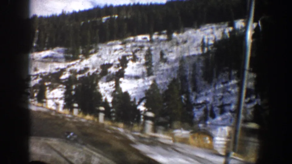 1961 Aspen, Colorado frame with a curving road, snow-covered hillside, evergreens, a small cabin, and a roadside utility pole.