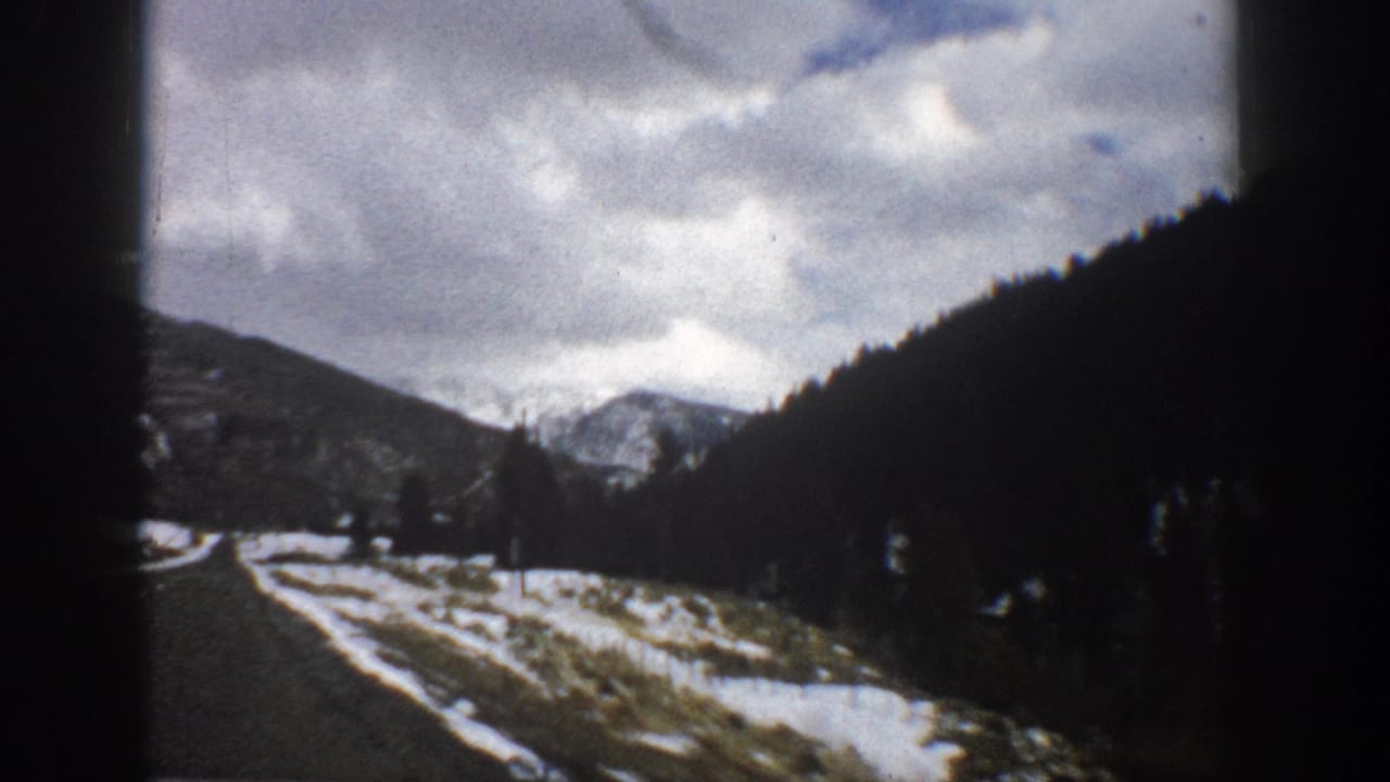Paved mountain road running through snow patches toward dark forested slopes.