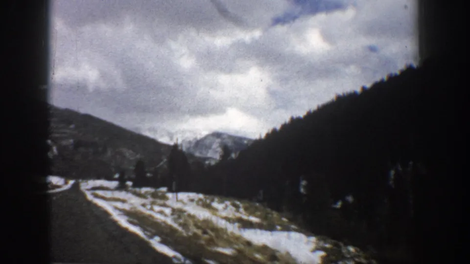 1961 Aspen, Colorado road-view frame with a paved lane, snow along the shoulders, forested hills, cloudy sky, and distant mountains ahead.