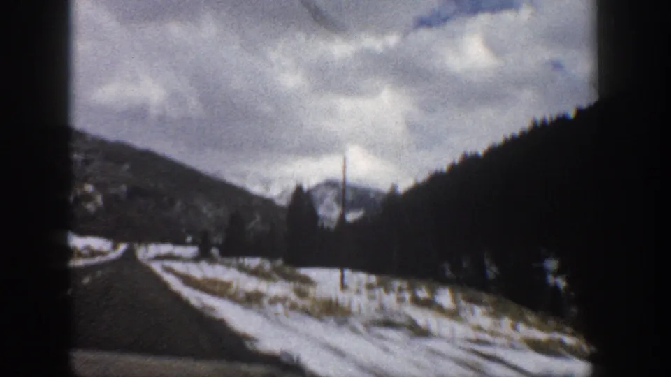 1961 Aspen, Colorado 8mm frame from a moving car, showing a paved road, snowy shoulder, dark tree-covered slope, and distant mountain ridge beneath clouds.