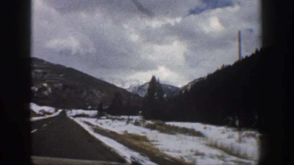 1961 Aspen, Colorado home-movie frame showing a paved road curving past patchy snow, evergreen slopes, roadside poles, and distant mountain peaks.