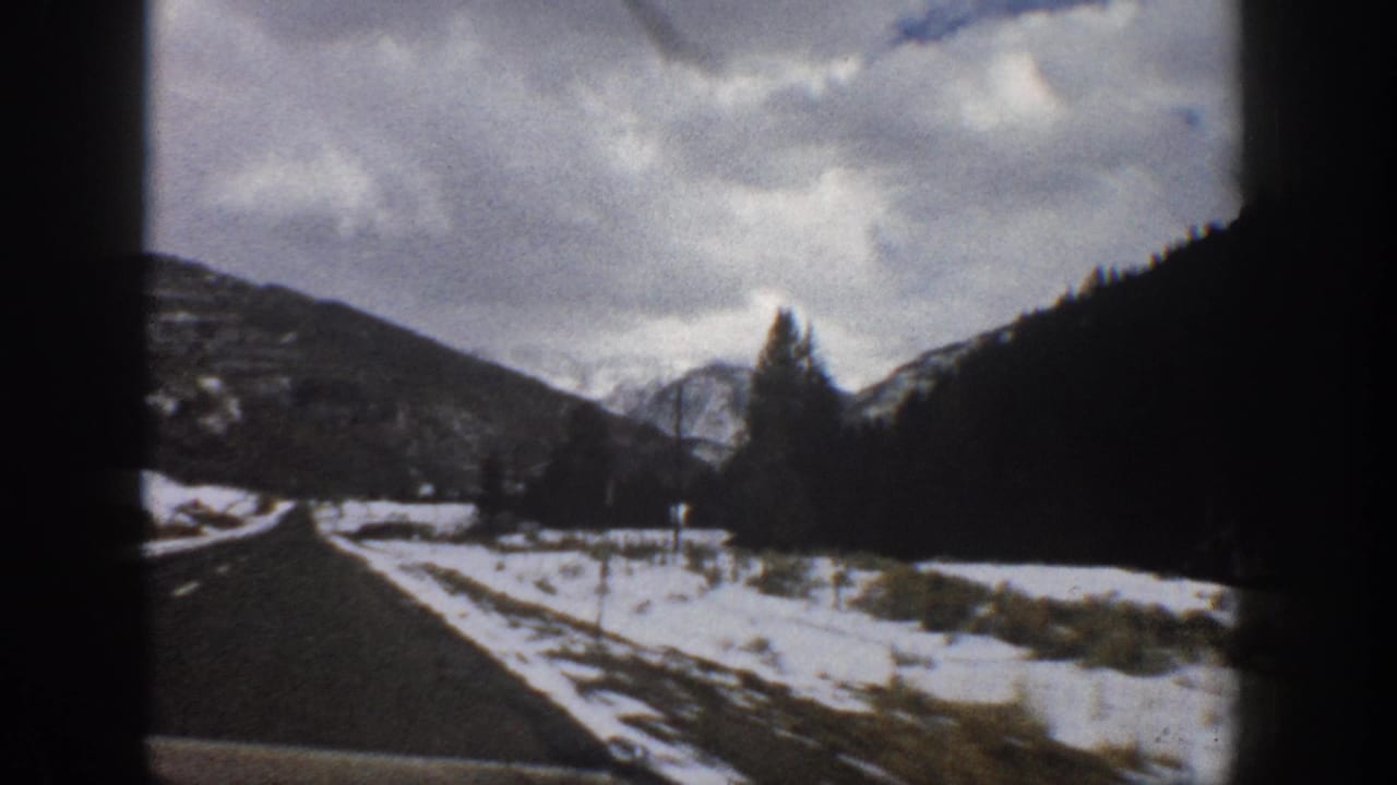 1961 Aspen, Colorado 8mm frame showing a road beside snow-covered ground, dark hills, evergreen trees, and distant mountains.