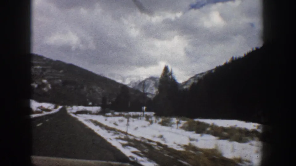 1961 Aspen, Colorado 8mm frame showing a road beside snow-covered ground, dark hills, evergreen trees, and distant mountains.