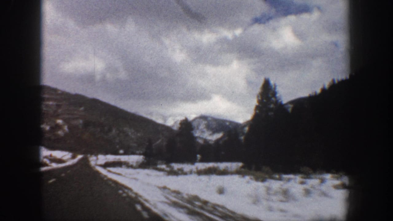 1961 Aspen, Colorado road-view frame with snow along the shoulder, evergreen trees, shadowed hills, and pale mountain peaks in the distance.