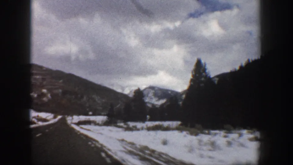 1961 Aspen, Colorado road-view frame with snow along the shoulder, evergreen trees, shadowed hills, and pale mountain peaks in the distance.