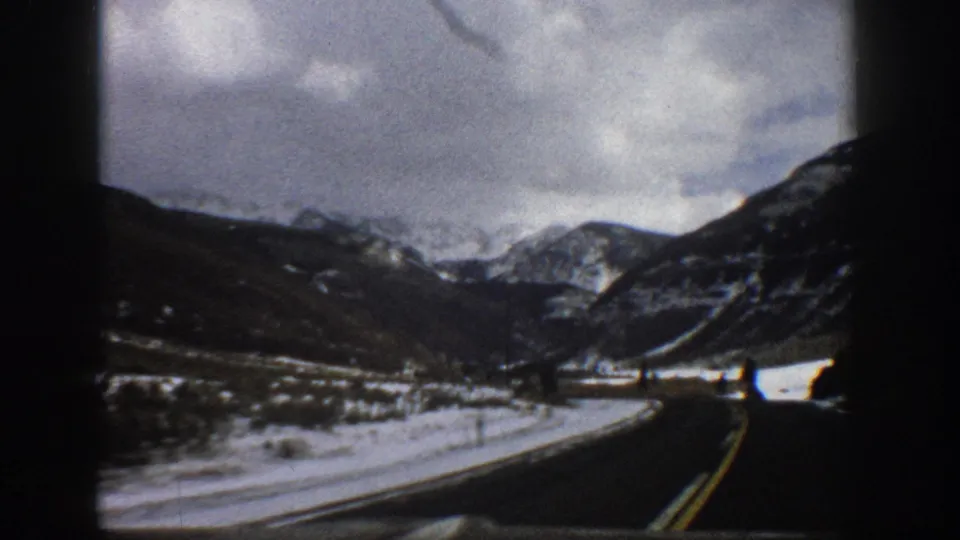 1961 Aspen, Colorado 8mm frame from a vehicle on a paved road approaching a broad mountain view with snow at the roadside.