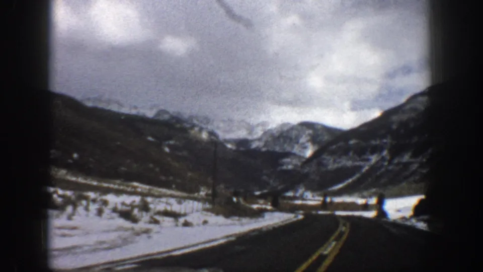1961 Aspen, Colorado 8mm frame showing a winding mountain road bordered by snow, open valley ground, dark slopes, and clouded peaks.