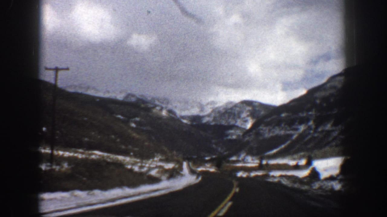 Curving road through a snowy valley with telephone poles, mountains, and heavy clouds.