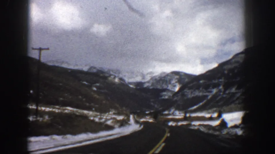 1961 Colorado mountain driving view with a curving paved road, snowfields, utility poles, and rugged peaks ahead.
