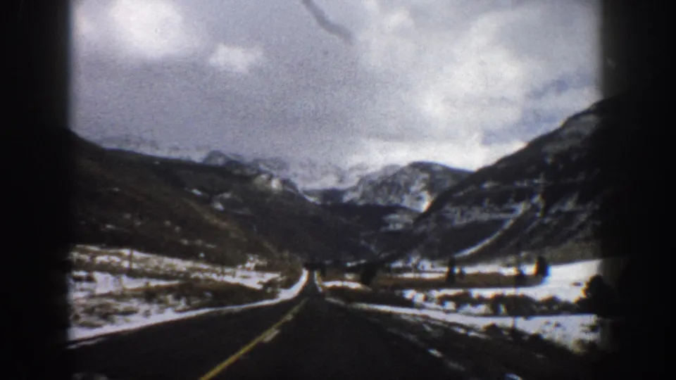 1961 Aspen-area 8mm frame looking down a two-lane road through snow toward rugged mountains under broken clouds.