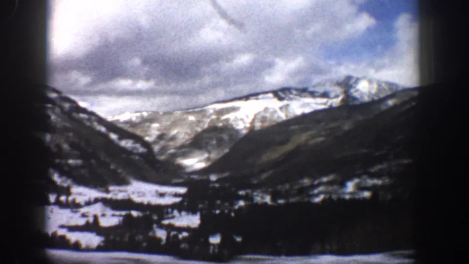 1961 Aspen-area 8mm frame showing snowy mountain ridges above a dark valley floor and scattered small structures or trees.