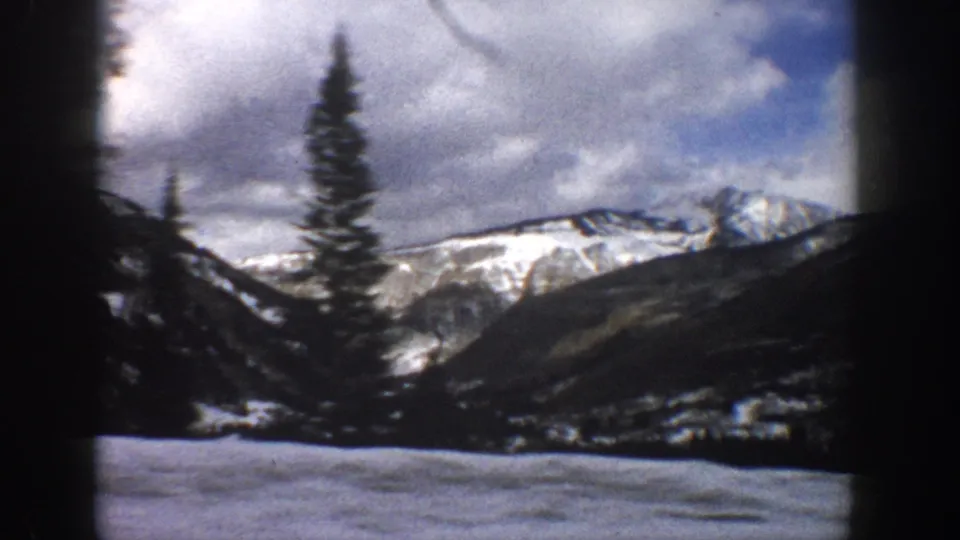 1961 Aspen-area 8mm frame with a snowy foreground, tall evergreens, and mountain ridges under a clouded sky.