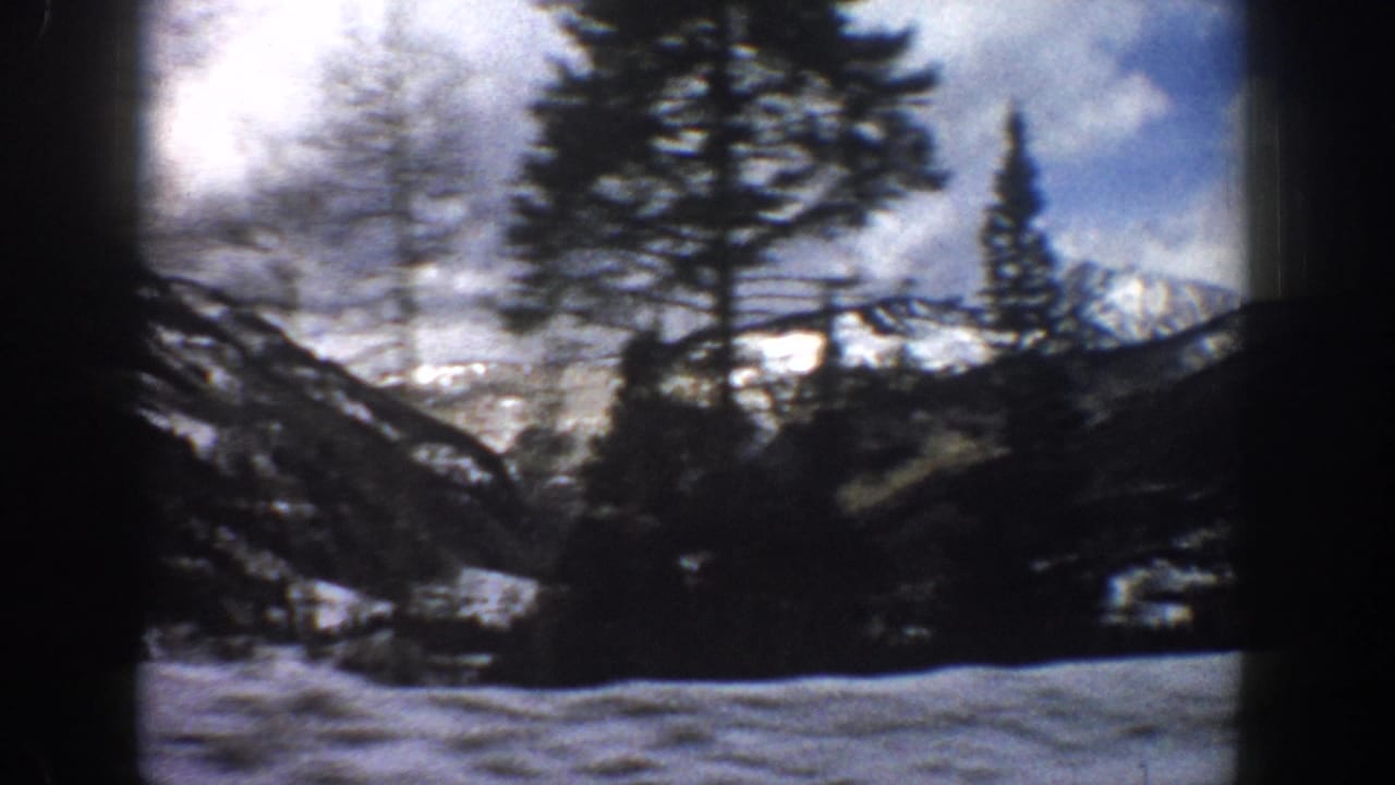 1961 Aspen-area 8mm frame showing snow, evergreens, bare trees, distant peaks, and a partly cloudy blue sky.