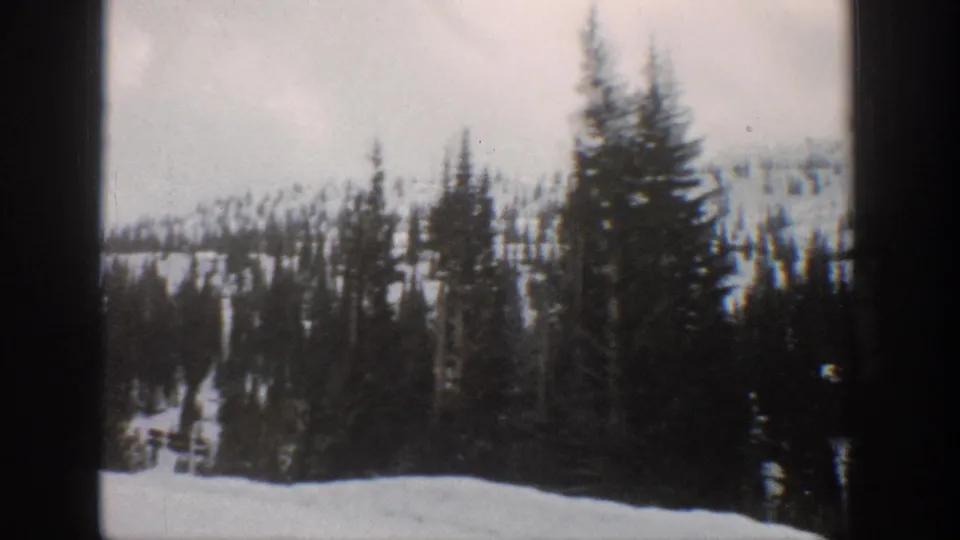 1961 Aspen-area 8mm frame showing snowbanks, evergreen trees, and a wooded mountain slope under an overcast sky.