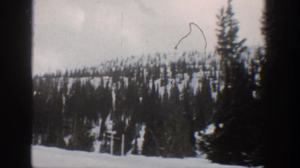 1961 Aspen-area home-movie frame with dark conifers, snowy hillside, pale sky, and visible film wear.