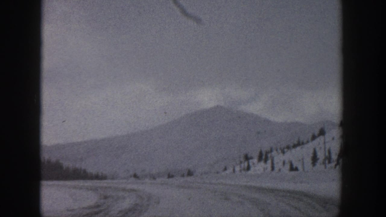 1961 Aspen, Colorado travel frame with a snowy road, distant mountain form, scattered trees, and muted winter light.
