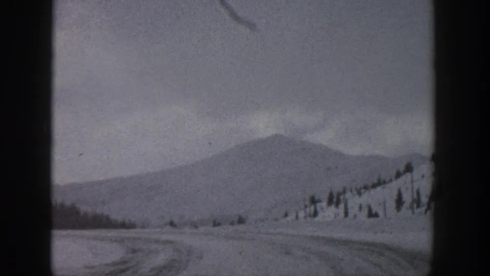 1961 Aspen, Colorado travel frame with a snowy road, distant mountain form, scattered trees, and muted winter light.