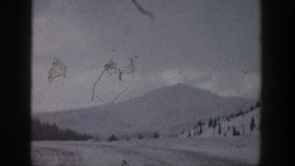 1961 Aspen, Colorado frame showing a hazy snowy road scene with evergreens, mountain slope, overcast sky, and visible film wear.