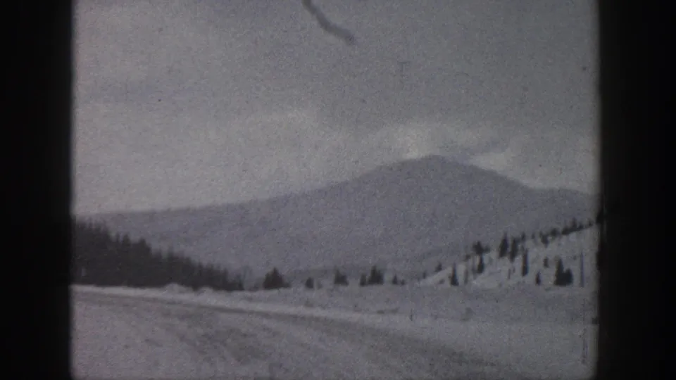1961 Aspen, Colorado 8mm frame of a snow-covered mountain road, hillside evergreens, cloudy sky, and soft monochrome exposure.