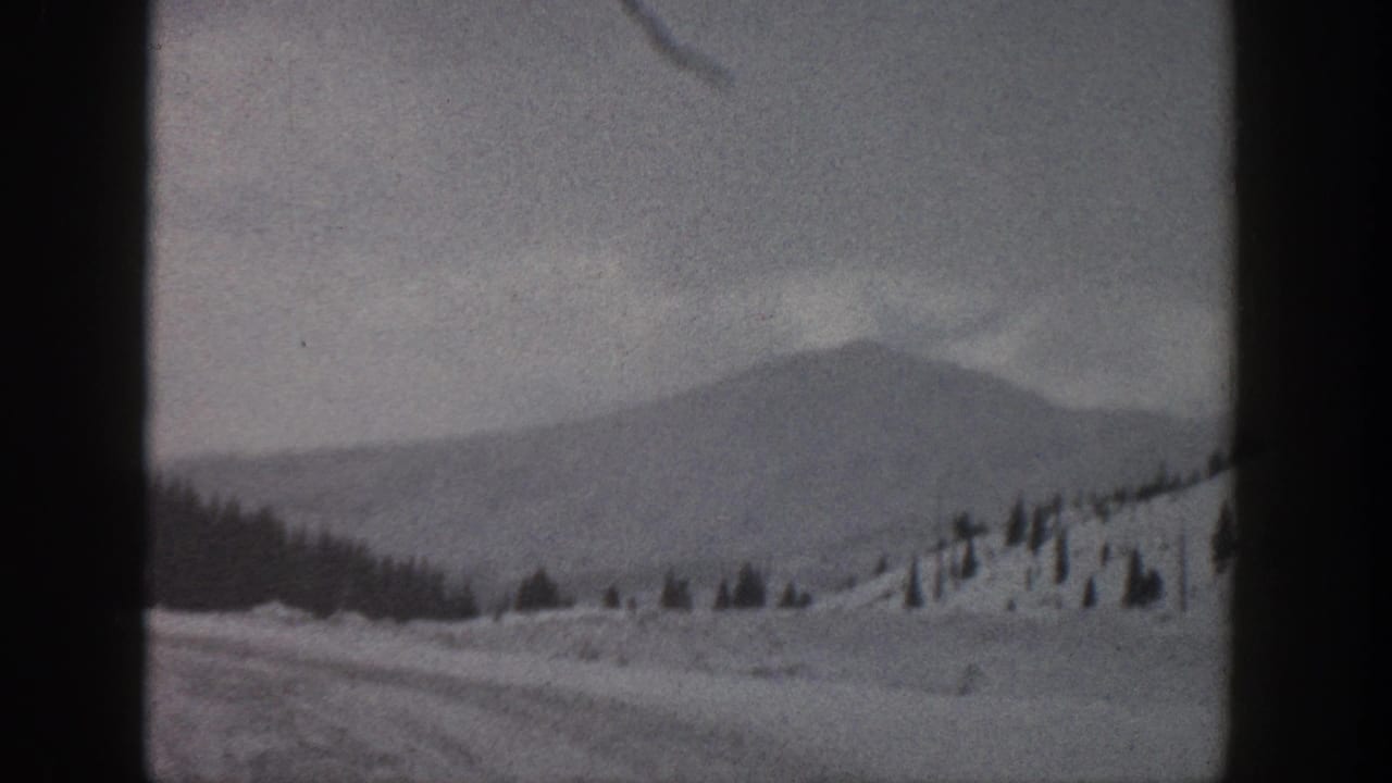 1961 Aspen, Colorado home-movie frame showing a snow-covered mountain slope, distant ridge, scattered evergreens, and dark 8mm frame edges.