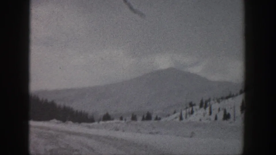 1961 Aspen, Colorado home-movie frame showing a snow-covered mountain slope, distant ridge, scattered evergreens, and dark 8mm frame edges.