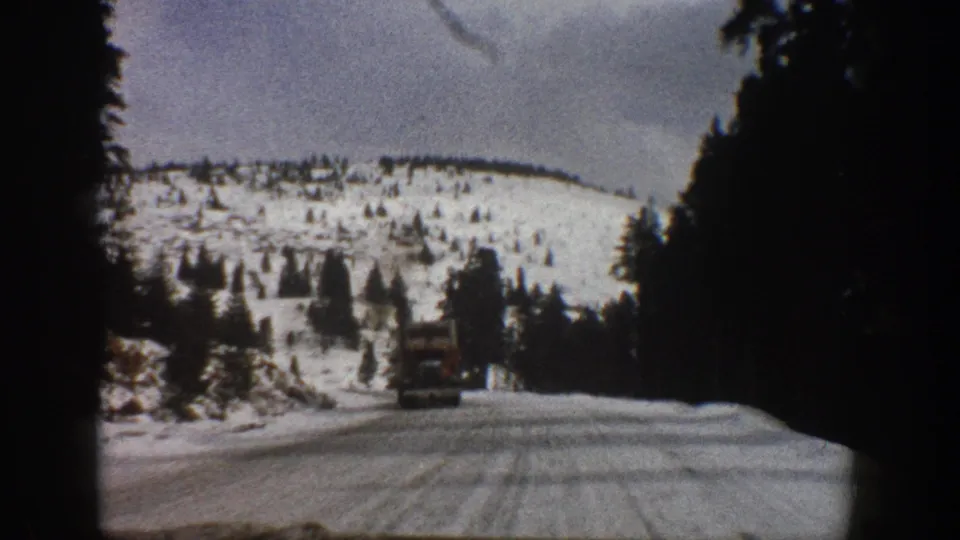 1961 Aspen-area 8mm frame showing a truck on a snowy mountain road bordered by evergreens and hillside snow.