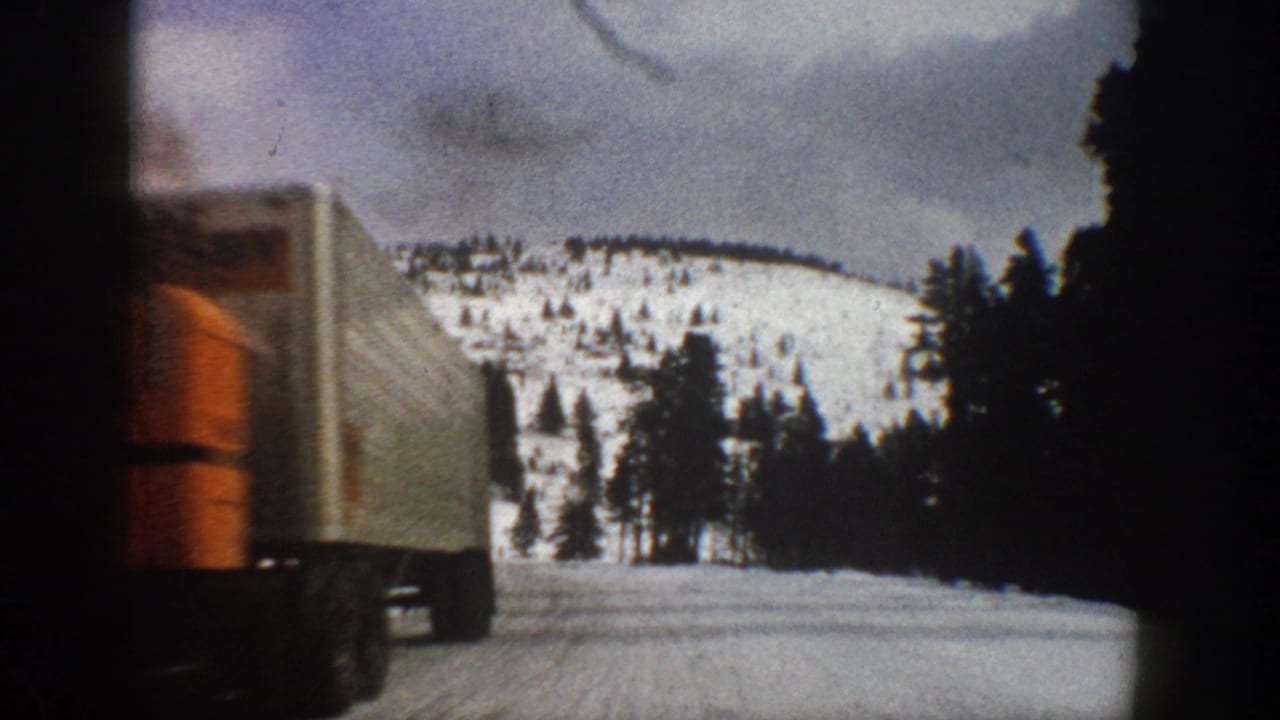 1961 Aspen, Colorado frame with a large orange-and-white semi-truck passing the camera on a snow-covered mountain road.