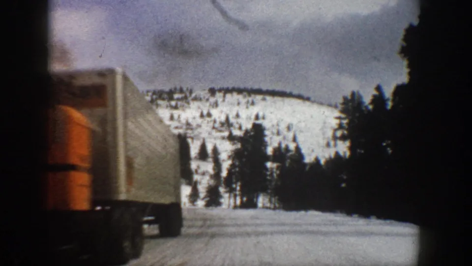 1961 Aspen, Colorado frame with a large orange-and-white semi-truck passing the camera on a snow-covered mountain road.