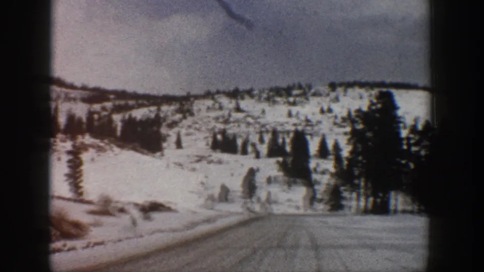1961 Aspen, Colorado frame showing a snowy mountain road curving past conifers with a white hillside beyond.