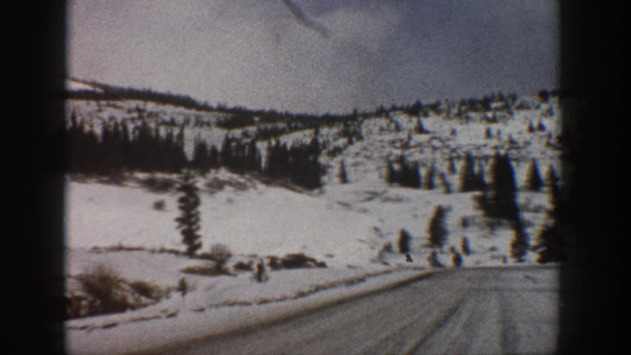1961 Aspen-area 8mm frame showing a snow-covered road ahead, open snowy hillsides, and scattered evergreens under a pale sky.