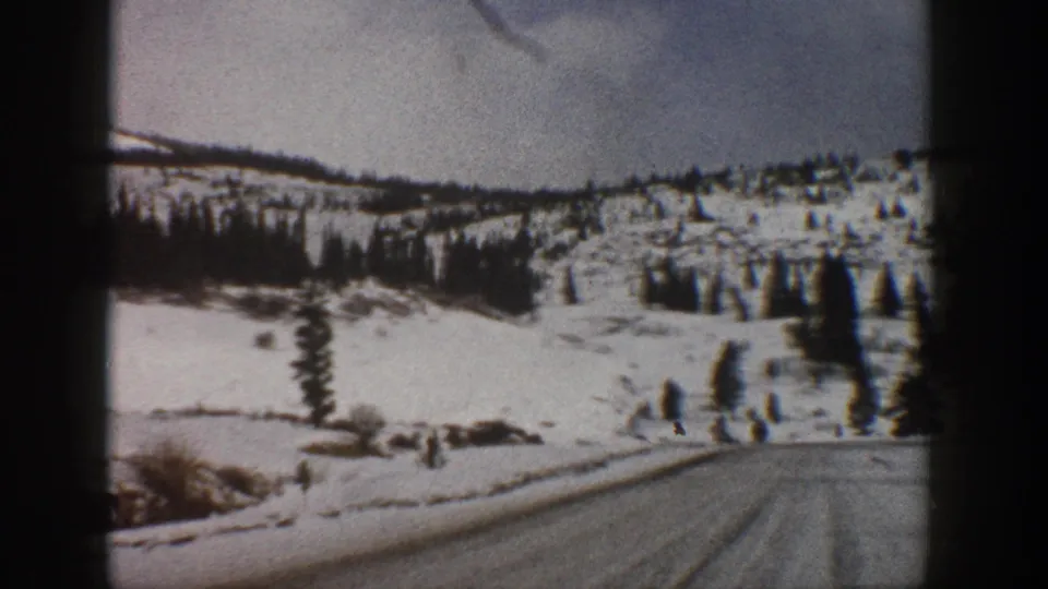 1961 Aspen-area 8mm frame showing a snow-covered road ahead, open snowy hillsides, and scattered evergreens under a pale sky.
