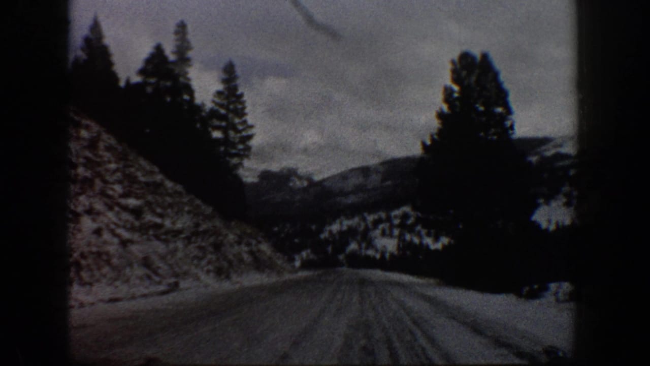 1961 Aspen-area 8mm frame from a moving vehicle on a snow-covered mountain road bordered by a rocky slope and evergreens.