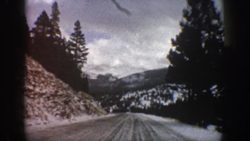 1961 Aspen-area 8mm frame showing a winter road with tire tracks, tall evergreens, a rocky bank, and snow-dusted hills beyond.