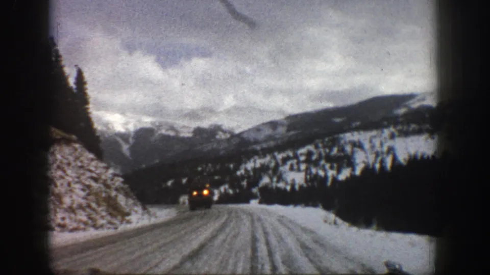 1961 Aspen-area 8mm frame showing a snow-covered road, mountain slopes, evergreens, and a distant vehicle with headlights visible.